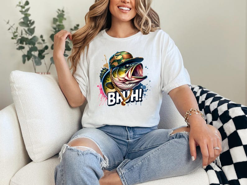 May include: White t-shirt featuring a colorful graphic of a bass wearing a camouflage hat and gold chain, with the word "BRUH!" in bold letters. The design includes splashes of pink, blue, and yellow.
