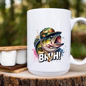 May include: White ceramic mug featuring a cartoon bass wearing a camouflage hat and gold chain. The fish has an open mouth and the word "BRUH!" is printed below. A s'more is on a wooden surface.