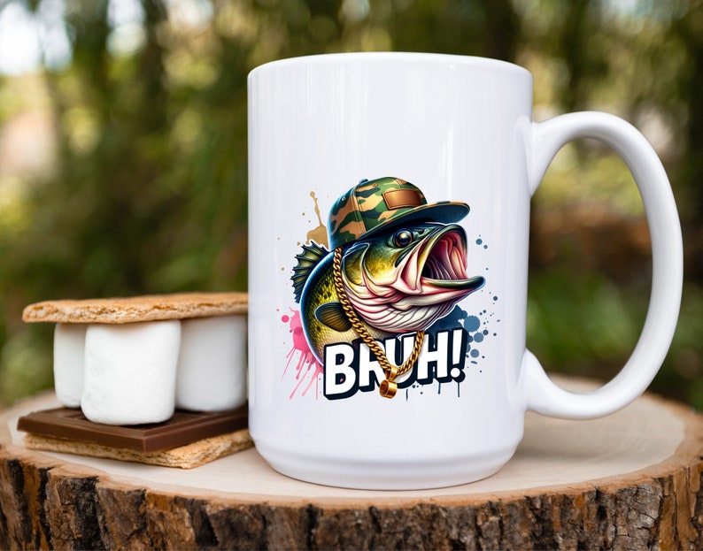 May include: White ceramic mug featuring a cartoon bass wearing a camouflage hat and gold chain. The fish has an open mouth and the word "BRUH!" is printed below. A s'more is on a wooden surface.