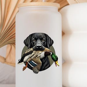 Pu&ograve; includere: Un bicchiere di vetro smerigliato con coperchio in legno e cannuccia. Il bicchiere presenta una grafica di un Labrador Retriever nero che tiene un'anatra in bocca, su un cerchio verde. L'anatra ha piume marroni, bianche e verdi.