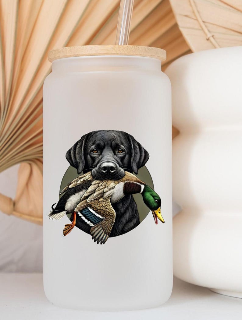 Pu&ograve; includere: Un bicchiere di vetro smerigliato con coperchio in legno e cannuccia. Il bicchiere presenta una grafica di un Labrador Retriever nero che tiene un'anatra in bocca, su un cerchio verde. L'anatra ha piume marroni, bianche e verdi.