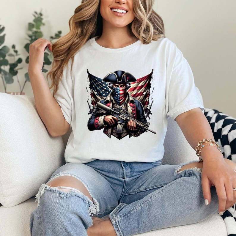 May include: White t-shirt with a graphic of a person wearing a tri-corner hat and bandana, holding a rifle, with an American flag behind them.