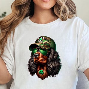 May include: White t-shirt with a graphic of a brown and black Cocker Spaniel wearing a camouflage baseball cap with the text "Lucky Dog" and green sunglasses. The dog is also wearing a green shamrock.