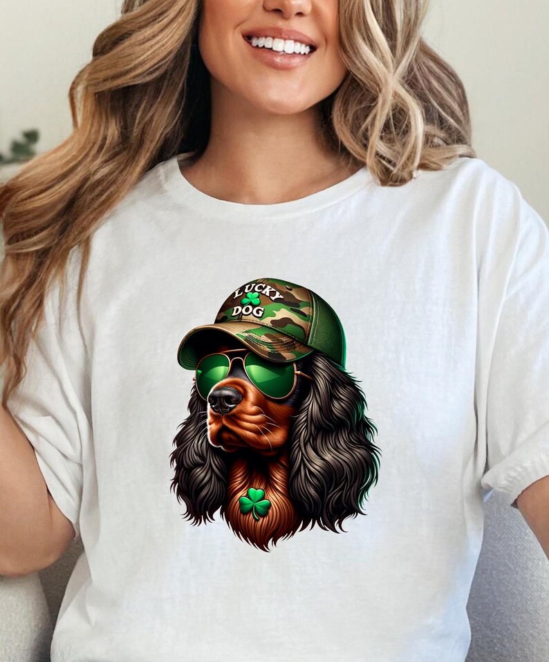 May include: White t-shirt with a graphic of a brown and black Cocker Spaniel wearing a camouflage baseball cap with the text "Lucky Dog" and green sunglasses. The dog is also wearing a green shamrock.