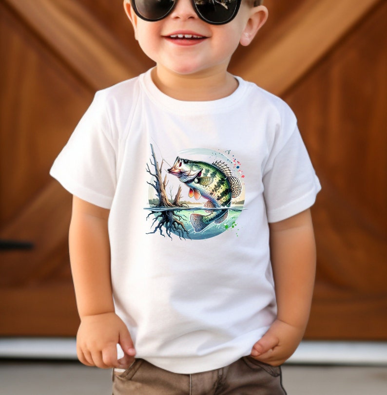 May include: White toddler t-shirt with a colorful graphic of a bass fish jumping out of the water. The fish is green, brown, and black with a white belly. The background is a blue circle with white and green splashes of paint.