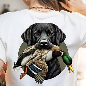 Pu&ograve; includere: T-shirt bianca con un disegno di un Labrador Retriever nero che tiene un'anatra. L'anatra ha piume marroni, bianche e verdi, su uno sfondo verde.