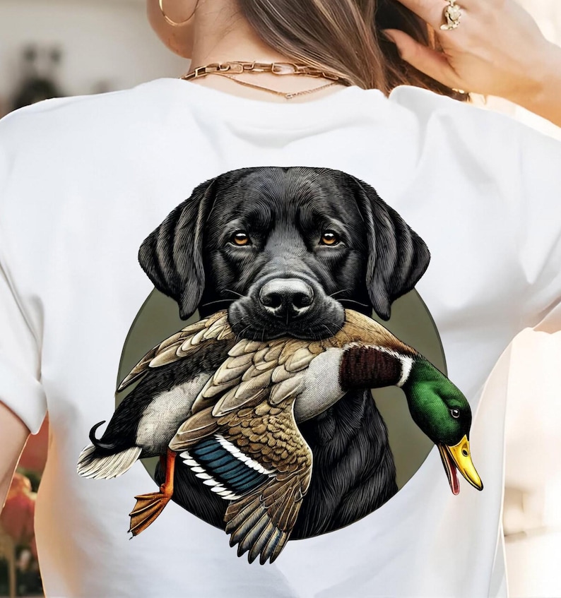 Pu&ograve; includere: T-shirt bianca con un disegno di un Labrador Retriever nero che tiene un'anatra. L'anatra ha piume marroni, bianche e verdi, su uno sfondo verde.