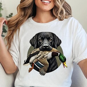 Pu&ograve; includere: T-shirt bianca con un disegno di un Labrador Retriever nero che tiene un'anatra. Il cane &egrave; posto su un cerchio verde. L'anatra ha piume marroni, bianche, verdi e arancioni.