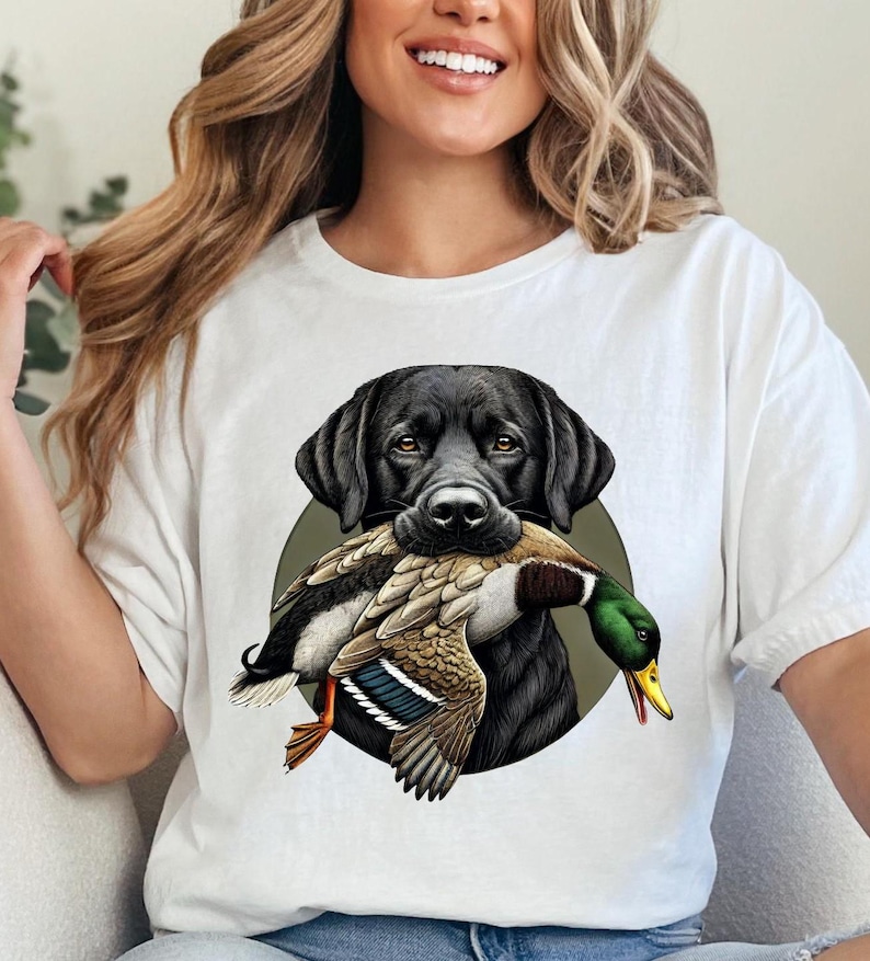 Pu&ograve; includere: T-shirt bianca con un disegno di un Labrador Retriever nero che tiene un'anatra. Il cane &egrave; posto su un cerchio verde. L'anatra ha piume marroni, bianche, verdi e arancioni.