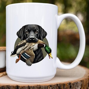 Pu&ograve; includere: Tazza in ceramica bianca con un'illustrazione di un Labrador Retriever nero che tiene un'anatra selvatica in bocca. L'anatra ha piume marroni, bianche e verdi. La tazza ha un manico grande ed &egrave; su uno sfondo verde sfocato.