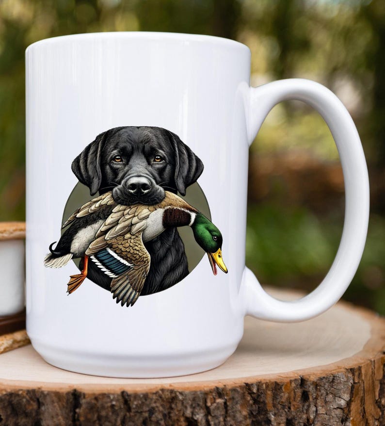 Pu&ograve; includere: Tazza in ceramica bianca con un'illustrazione di un Labrador Retriever nero che tiene un'anatra selvatica in bocca. L'anatra ha piume marroni, bianche e verdi. La tazza ha un manico grande ed &egrave; su uno sfondo verde sfocato.