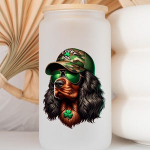 May include: A white frosted glass water bottle with a bamboo lid and a straw. The bottle has a graphic of a brown and black dog wearing a green camouflage hat with the words "Lucky Dog" on it. The dog is wearing green sunglasses and has a green shamrock around its neck.