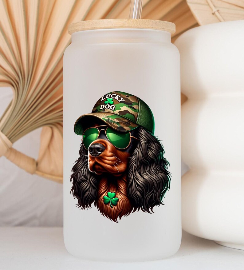May include: A white frosted glass water bottle with a bamboo lid and a straw. The bottle has a graphic of a brown and black dog wearing a green camouflage hat with the words "Lucky Dog" on it. The dog is wearing green sunglasses and has a green shamrock around its neck.