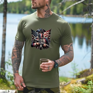 May include: A green t-shirt with a graphic of a person wearing a bandana and a tri-cornered hat holding a rifle. The person is standing in front of an American flag.