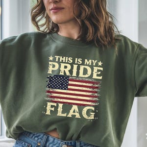 Puede incluir: Una sudadera verde con un gr&aacute;fico de bandera estadounidense desgastado y el texto "This is my pride flag".