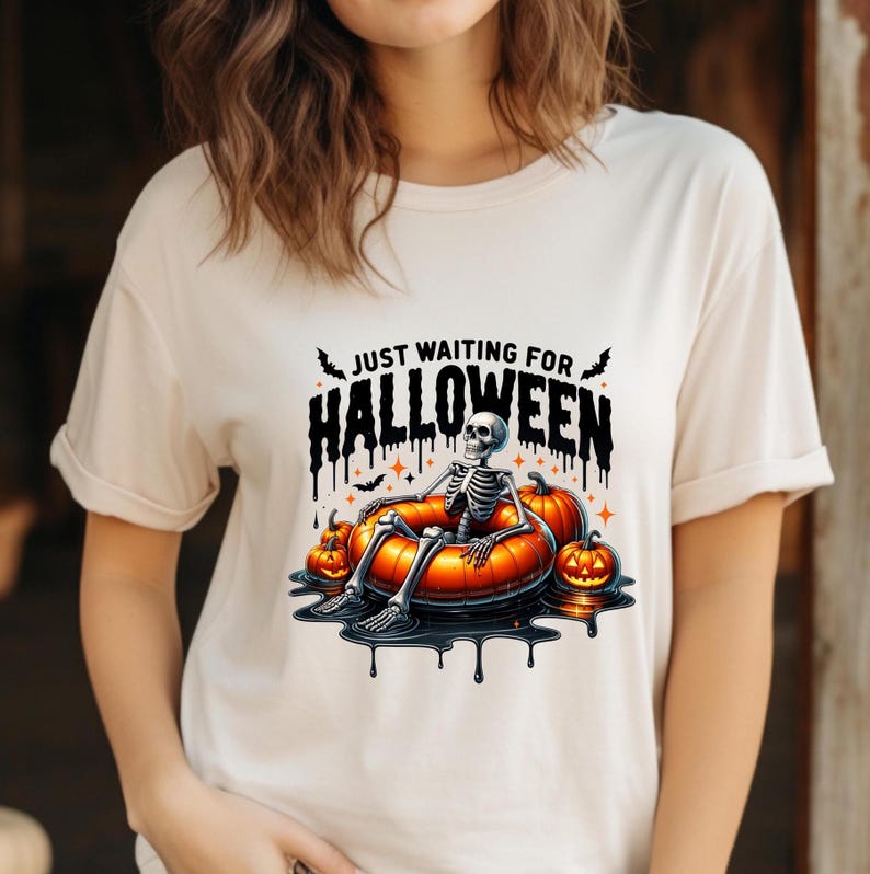 May include: A white t-shirt with the text "Just Waiting for Halloween" in black and dripping letters. A skeleton is sitting in a pumpkin-shaped inner tube surrounded by pumpkins.