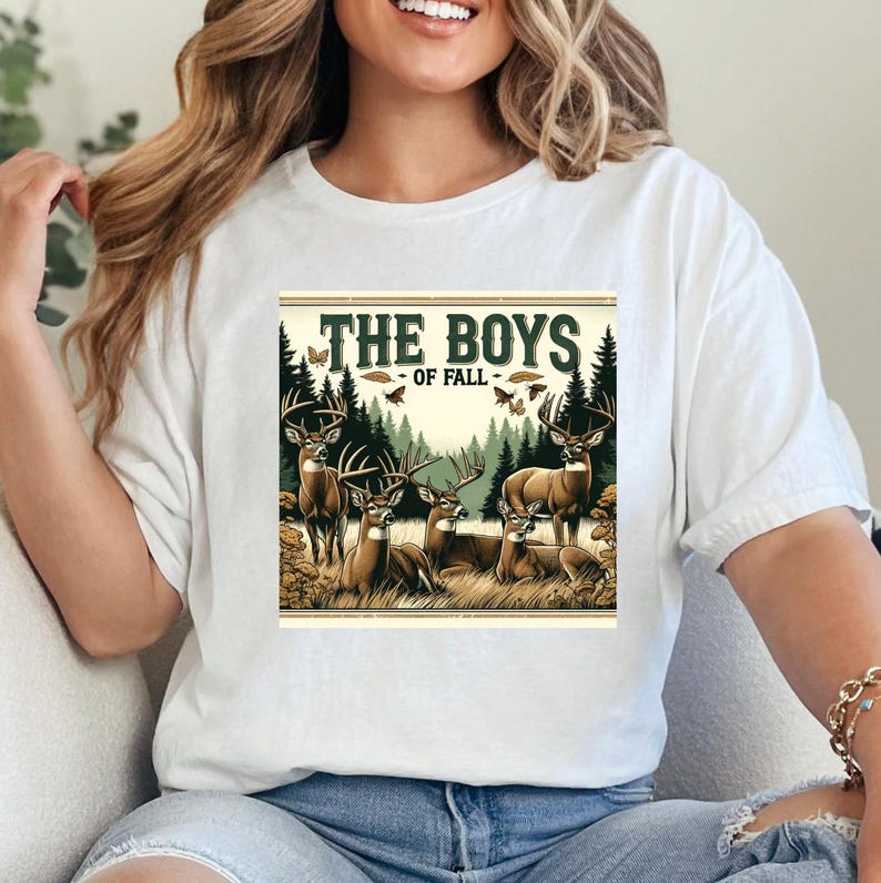 K&ouml;nnte beinhalten: Wei&szlig;es T-Shirt mit einem grafischen Druck einer Gruppe von Hirschen in einer Waldlandschaft. Der Text "THE BOYS - OF FALL -" ist &uuml;ber den Hirschen gedruckt.