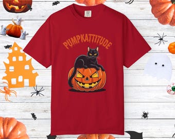 May include: A red t-shirt with a Halloween design. It features a black cat perched on an orange pumpkin with a carved face. The text "PUMPKATTITUDE" is above the cat.