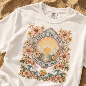 以下が含まれることがあります： 花と海のグラフィックがプリントされた白いTシャツ。デザインは夕日、波、そして「CHASE THE SUN LIVE IN THE LIGHT」のテキストが特徴です。柔らかい素材で作られており、クルーネックです。
