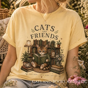 Peut inclure: T-shirt jaune clair avec l'inscription "CATS ARE FRIENDS" au-dessus d'une illustration de chat lisant un livre devant une bibliothèque. Le t-shirt porte également l'inscription "READ MORE • GROW MORE • BE KIND" et "CATS AND BOOKS AESTHETIC".