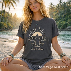 Peut inclure: T-shirt gris foncé avec un motif doré représentant une personne méditant devant un soleil et des vagues, avec l'inscription "rise & align". L'image est étiquetée "Soft Yoga aesthetic".
