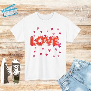 Peut inclure: T-shirt blanc avec le mot "LOVE" en lettres rouges et roses avec un contour blanc. Les lettres sont entourées de petits cœurs roses et rouges.