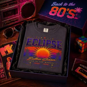 Peut inclure: Un t-shirt gris foncé avec le mot "ECLIPSE" en violet et orange, avec un graphisme de coucher de soleil et le texte "Endless Summer". Le t-shirt est présenté dans un coffret cadeau sur le thème rétro des années 80, avec un lecteur de cassettes et des lunettes de soleil.