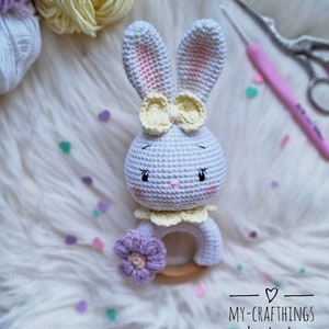 May include: White crocheted bunny rattle with a yellow bow and a purple flower. The rattle has a wooden ring and is handmade.