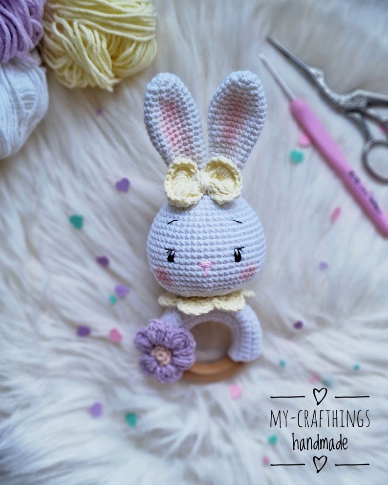 May include: White crocheted bunny rattle with a yellow bow and a purple flower. The rattle has a wooden ring and is handmade.