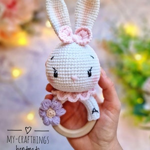 May include: A crocheted bunny rattle with a pink bow, a purple flower, and a wooden ring. The rattle is white with pink accents and has a letter "A" embroidered on the ring.