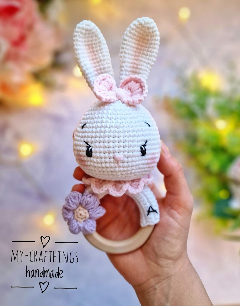 May include: A crocheted bunny rattle with a pink bow, a purple flower, and a wooden ring. The rattle is white with pink accents and has a letter "A" embroidered on the ring.