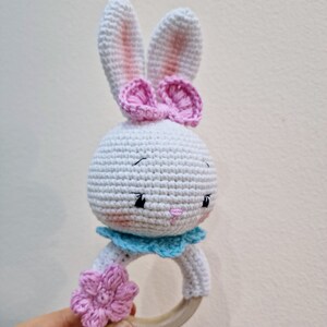 May include: White crocheted bunny rattle with pink bow and flower accents. The rattle has a wooden ring.