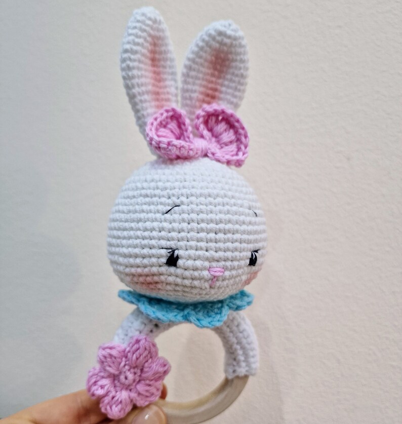 May include: White crocheted bunny rattle with pink bow and flower accents. The rattle has a wooden ring.