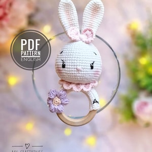 May include: A crocheted white bunny rattle with pink ears and a pink bow. The bunny is attached to a wooden ring with a purple flower.