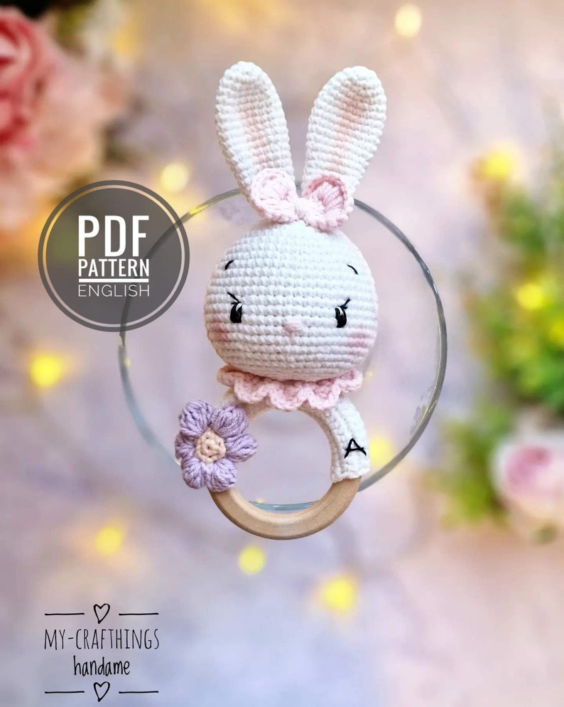 May include: A crocheted white bunny rattle with pink ears and a pink bow. The bunny is attached to a wooden ring with a purple flower.
