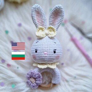 May include: A white crocheted bunny rattle toy with a yellow bow, purple flower, and wooden ring. The bunny has pink cheeks and black eyes.