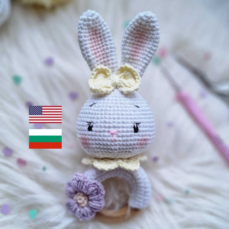 May include: A white crocheted bunny rattle toy with a yellow bow, purple flower, and wooden ring. The bunny has pink cheeks and black eyes.