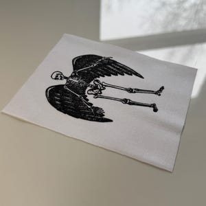 May include: A white square patch with a black illustration of a winged skeleton. The skeleton is depicted lying down with its legs extended. The wings are detailed with multiple feathers.