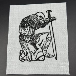 May include: Black and white embroidered patch depicting a kneeling knight holding a sword. The knight is wearing armour and a long robe.