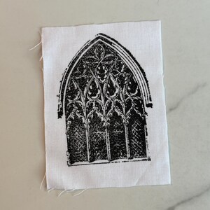 May include: A black and white fabric patch featuring a gothic arched window design. The window is intricately detailed with floral and geometric patterns. The patch is rectangular with raw edges, and the design is printed on a white fabric.
