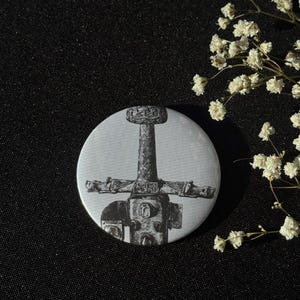 Könnte beinhalten: Ein runder, weißer Button mit einer detaillierten Schwarz-Weiß-Illustration eines Schwertes. Das Schwert hat einen kreuzförmigen Knauf und einen strukturierten Griff. Der Button befindet sich vor einem dunklen Hintergrund mit weißen Blüten.