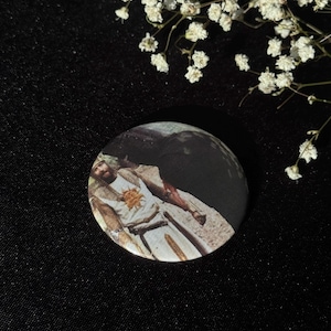 Op de afbeelding: Ronde knoop met een afgedrukte afbeelding van een persoon in een witte tuniek met een zonnembleem. De knoop staat tegen een zwarte achtergrond met witte bloemen.