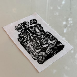 May include: A small, rectangular patch featuring a black and white illustration of a knight on horseback in combat. The image is printed on white fabric with frayed edges, giving a vintage or handmade feel.