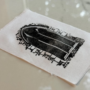 Könnte beinhalten: Ein kleines, rechteckiges Stoffstück in Weiß mit einer schwarzen Bootsillustration. Das Boot ist in einem Linolschnittstil dargestellt, mit Details, die ein Ruderboot oder ein ähnliches Schiff andeuten. Die Stoffkanten sind roh und unfertig.