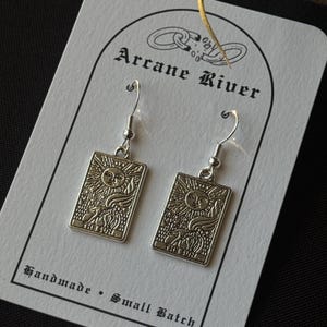 May include: Silver-toned earrings featuring a rectangular design with a sun motif, labeled "The Sun." The earrings hang from silver hooks and are displayed on a card with the text "Arcane River" and "Handmade • Small Batch."
