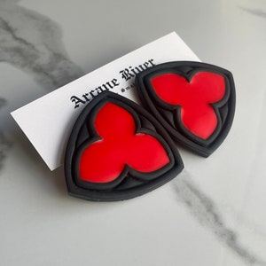 Può includere: Due orecchini neri e rossi con un design gotico. Ogni orecchino presenta una forma rossa a tre lobi all'interno di un bordo nero. Una carta bianca con il testo "Arcane River" è parzialmente visibile.