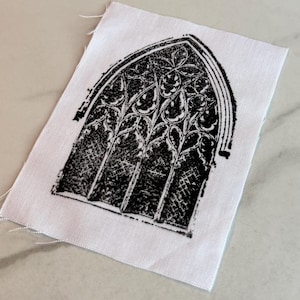 May include: A black and white fabric square featuring a gothic arched window design. The window is intricately detailed with floral and geometric patterns. The fabric has a raw edge.