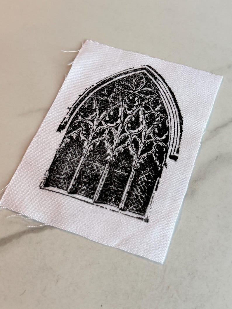 May include: A black and white fabric square featuring a gothic arched window design. The window is intricately detailed with floral and geometric patterns. The fabric has a raw edge.
