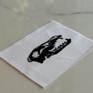 Pode incluir: Um remendo de tecido branco com uma imagem impressa em preto de uma caveira de animal.