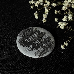 Könnte beinhalten: Ein runder, grauer Button mit dem Text "I SPEAK FOR THE TREES" in einer stilisierten Schriftart. Der Button zeigt eine Schwarz-Weiß-Illustration eines Baumes und einer Landschaft. Der Hintergrund ist schwarz, mit weißen Blumen in der oberen rechten Ecke.